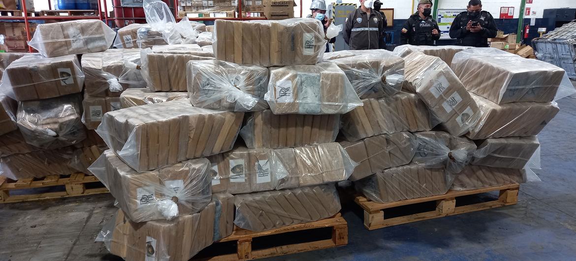 Large stacks of seized cocaine packages wrapped in plastic, with police officers observing in the background at a warehouse in Ecuador.